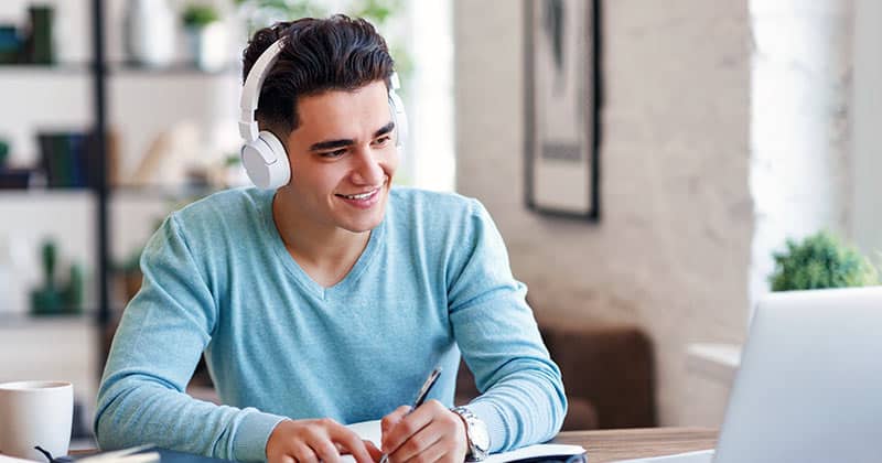 Male student with headphones taking notes off laptop